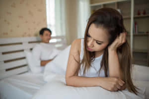 一位女士在床上看起來悲傷，而一位男士坐在她後面的臥室裡。 — 悲傷的愛情 壁紙