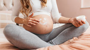 孕婦坐在床上，在肚子上塗抹乳液形成笑臉形狀。 — 懷孕 壁紙
