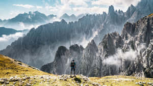 健行者站在山脊上，面對下方有雲層的戲劇性岩石山峰。 — 山脈 壁紙