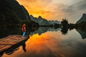 夕陽下，竹筏上站立的人，平靜水面倒映著山巒。 — 自然 壁紙
