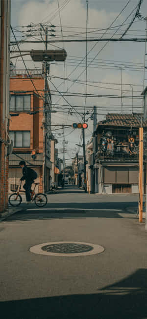 一個人在有建築物和電線的狹窄城市街道上騎自行車。 — iPhone 16e 壁紙