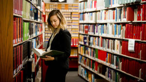 穿著黑色西裝外套的女性在圖書館書架之間閱讀書籍。 — 工作 壁紙