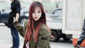 紅髮年輕女子在城市街道上揮手，穿著綠色外套 — K-pop 壁紙
