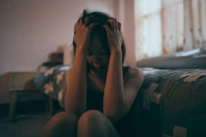 年輕女性坐在床上雙手抱頭神情痛苦在光線昏暗的房間 — 憂鬱 GIF 壁紙