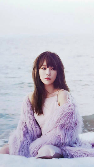 一位穿著蓬鬆紫色外套的女性，在寧靜的肖像中坐在海邊。 — 태연 壁紙