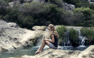 一位女性赤腳坐在森林地區小瀑布附近的岩石上。