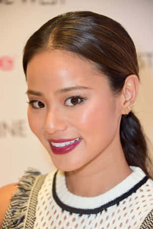 深色頭髮紮成馬尾的女性對著鏡頭微笑，身穿白色針織上衣搭配黑色飾邊和紅色唇膏。 — 凱莉·珍娜 壁紙