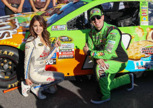 NASCAR車手和模型在陽光明媚的日子裡在一輛色彩繽紛的賽車旁擺姿勢。 — 運動 壁紙