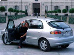 女士從宏偉建築前的銀色汽車打開車門中揮手。 — 高清汽車 壁紙