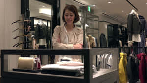 女性在服裝店展示櫃後微笑，櫃內有毛衣和手提包。 — 購物 壁紙