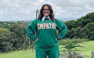 穿著綠色 'EMPATHY' 運動衫的女性在戶外長滿青草的山丘上微笑，背景有樹木和山脈。 — 克莉絲汀·史都華 壁紙
