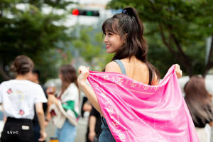 年輕女子微笑著在城市街道上舉起一條粉色圍巾。 — 平井もも 壁紙