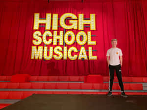 年輕男子在紅色劇院舞台上微笑，身後有「High School Musical」標誌。 — 迪士尼角色 壁紙