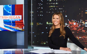 新聞主播 Trish Regan 在桌前微笑，身後是城市燈光。 — 福斯新聞 壁紙
