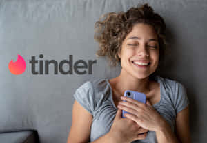 女性在灰色沙發上拿著手機微笑，Tinder 標誌可見 — 可愛的愛情 壁紙