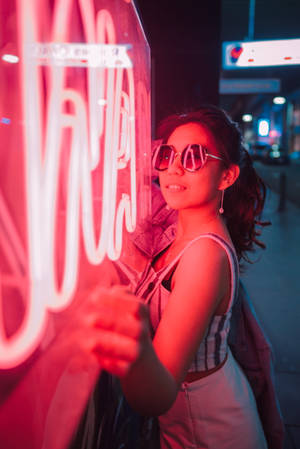 戴太陽眼鏡的女士在夜晚的城市街道上，在發光的紅色霓虹燈牌旁邊微笑。 — 霓虹紅 壁紙