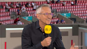 BBC Sport評論員在體育場中微笑著拿著麥克風。 — 16:9 壁紙
