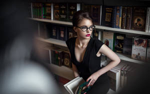 戴眼鏡的女性在圖書館裡拿著一本書，身後有書架。 — 看書 GIF 壁紙