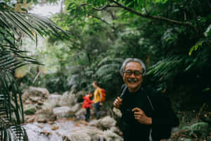 一位戴眼鏡和揹背包的長者，在鬱鬱蔥蔥的綠色森林中徒步旅行，背景有溪流和兩名徒步旅行者，他微笑著。 — 日本櫻花 壁紙