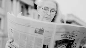 一位戴眼鏡的老年女性在戶外閱讀黑白報紙。 — Old Paper 壁紙