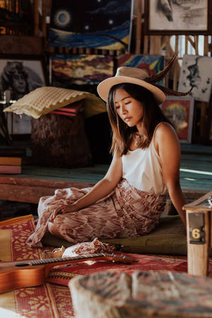 一位戴著羽毛帽子的女士，安靜地坐在充滿藝術品和吉他的創意工作室中。 — 曼谷 壁紙