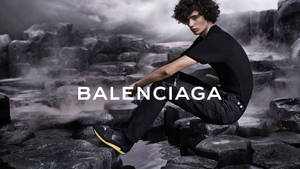 模特兒穿著 Balenciaga 服裝，坐在濕漉漉的火山岩上，背景霧氣瀰漫。 — 純黑 壁紙