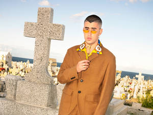 穿著米色外套和黃色太陽眼鏡的男士站在一個有海景的墓地十字架旁。 — 壞兔子 GIF 壁紙