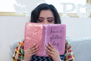 女性手持亮粉日記，上面有訊息 have a glitter day，坐在沙發上。 — 正向 壁紙