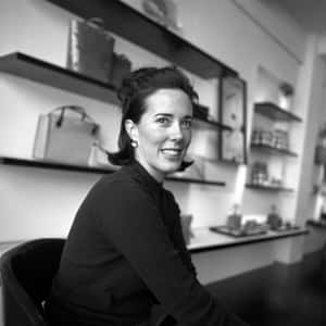一位女士在現代零售空間中微笑坐著，旁邊有陳列手袋的架子。 — Kate spade 壁紙