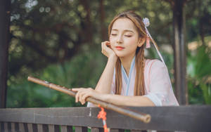 年輕女子穿著傳統粉紅色長袍，在寧靜的花園環境中手持竹笛。 — 模特兒 壁紙
