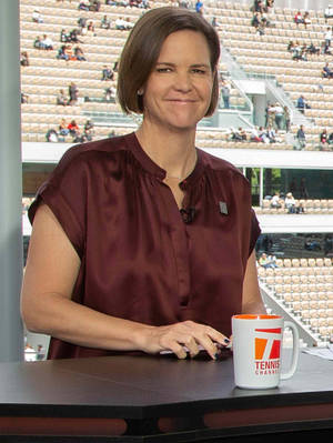 女士在辦公桌前微笑，手持Tennis Channel馬克杯，背景是體育場人群。 — 運動 壁紙