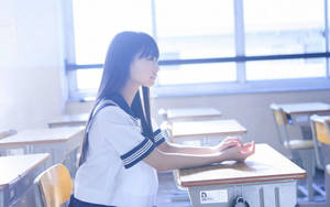 一位穿著水手服的學生坐在空教室的書桌前，望著窗外。 — 日本 壁紙