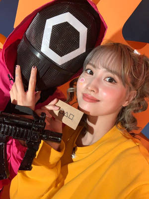 年輕女性穿著黃色連帽衫，手持面具和玩具槍，燦爛微笑。 — 067 魷魚遊戲 壁紙