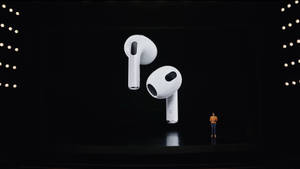 一個人在黑暗的舞台上展示帶有發光燈光的白色無線耳塞。 — AirPods 壁紙