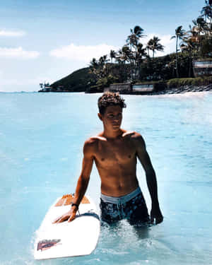 年輕男子在淺海中手持衝浪板，背景有棕櫚樹和小山丘。 — 4:5 壁紙