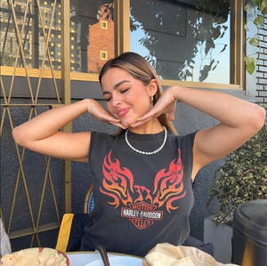 身穿Harley-Davidson T恤的女性在戶外咖啡館微笑，雙手框著臉部。 — 艾迪生·雷 壁紙