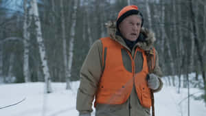年長獵人穿著橙色背心站在雪地樹林中，看起來很害怕。 — 湯姆·費爾頓 壁紙