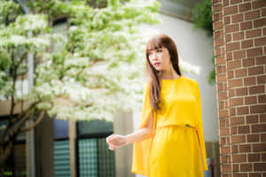 黃裙女性站在磚牆與開花樹木旁 — 黃色美學 GIF 壁紙