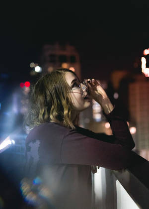 女性在夜晚的城市陽台上從玻璃杯飲水，背景模糊燈光。 — 夜景城市 壁紙