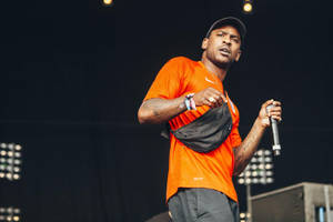 男性歌手在舞台上表演，身穿橙色襯衫和帽子，手持麥克風。 — Skepta 壁紙