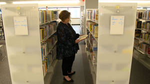 女性在白色書架之間的圖書館通道中瀏覽書籍。 — 工作 壁紙