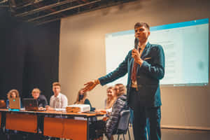 年輕男子在演講廳向帶有筆記型電腦的觀眾進行簡報。 — 海軍藍 壁紙
