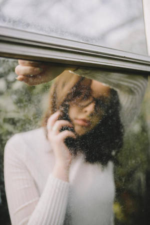 身穿白色毛衣的女性在雨痕斑斑的窗戶後打電話 — 女孩感手機 壁紙
