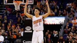 籃球運動員在比賽中高舉雙臂慶祝，面前是歡呼的球迷。 — Devin Booker 壁紙