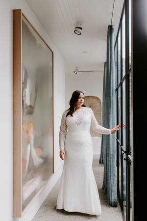 身穿白色蕾絲婚紗的女性站在現代走廊的玻璃門旁。 — 婚紗 壁紙