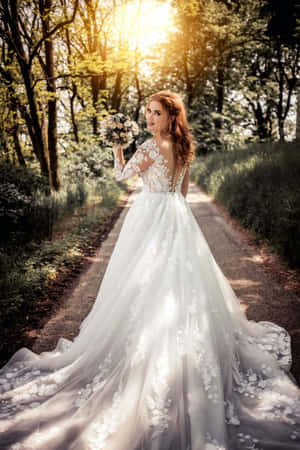 身穿白色蕾絲婚紗的新娘，手持花束，走在陽光明媚的森林中。 — 婚紗 壁紙