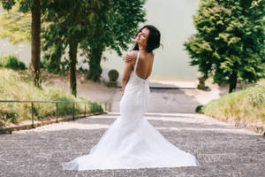 身穿白色蕾絲婚紗的新娘在綠樹環繞的花園石徑上微笑。 — 婚紗 壁紙