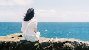 身穿白衣的女子坐在石牆上，俯瞰日落時分的海洋 — 海灘 壁紙