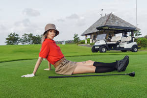 身穿紅色Polo衫和米色短褲的女性，在高爾夫球場的綠色草地上，旁邊有高爾夫球桿和球車，正在休息。
