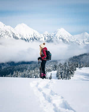 登山者手持登山杖、背著背包，站在白雪覆蓋的山頂上，眺望森林和雲層 — 冬季風景 壁紙